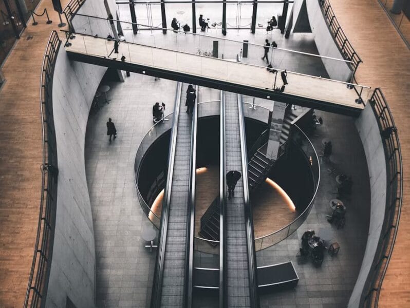 Vue en plongée d'un espace de bureau spacieux avec des escalators, un plafond en verre, et des personnes marchant sur un pont